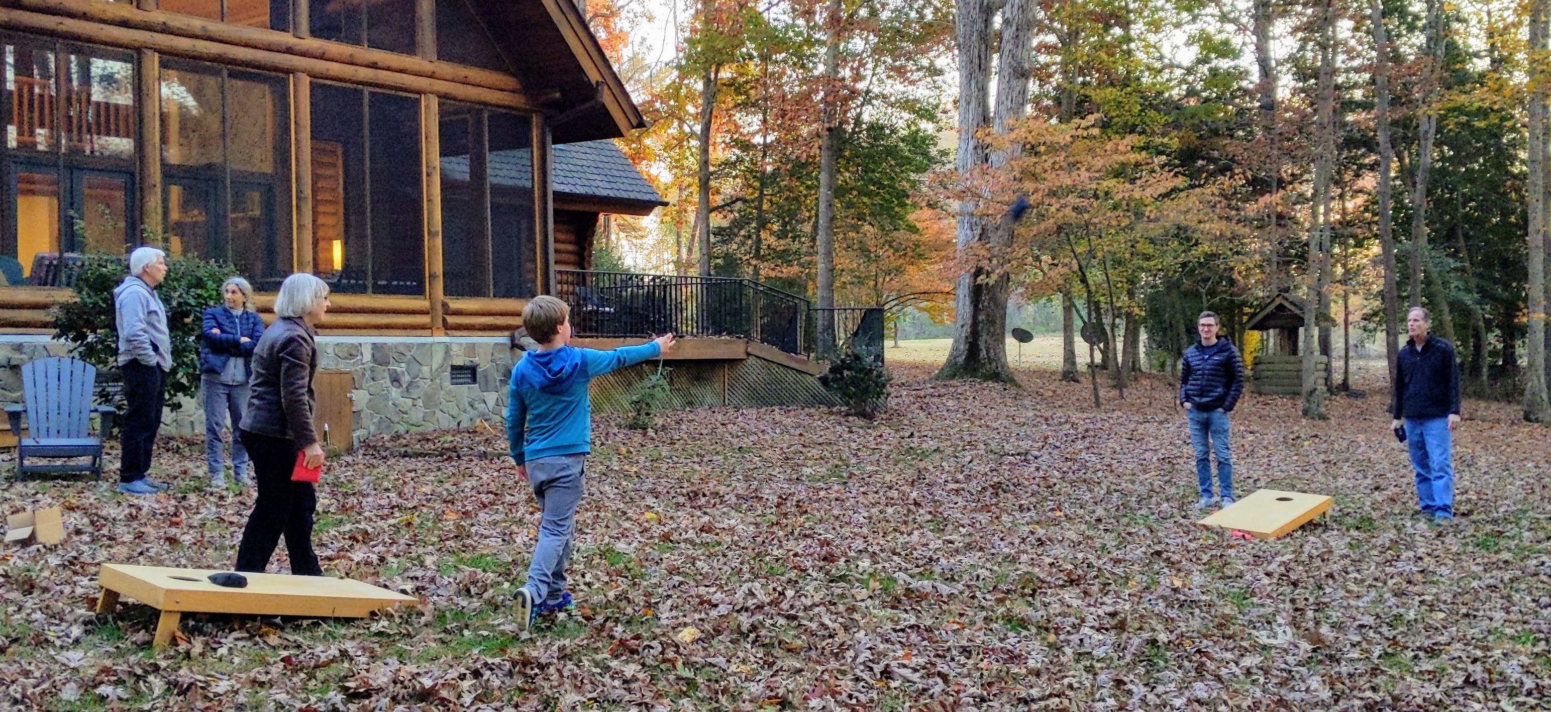 Four people playing corn hole  in the fall with others talking in the background.