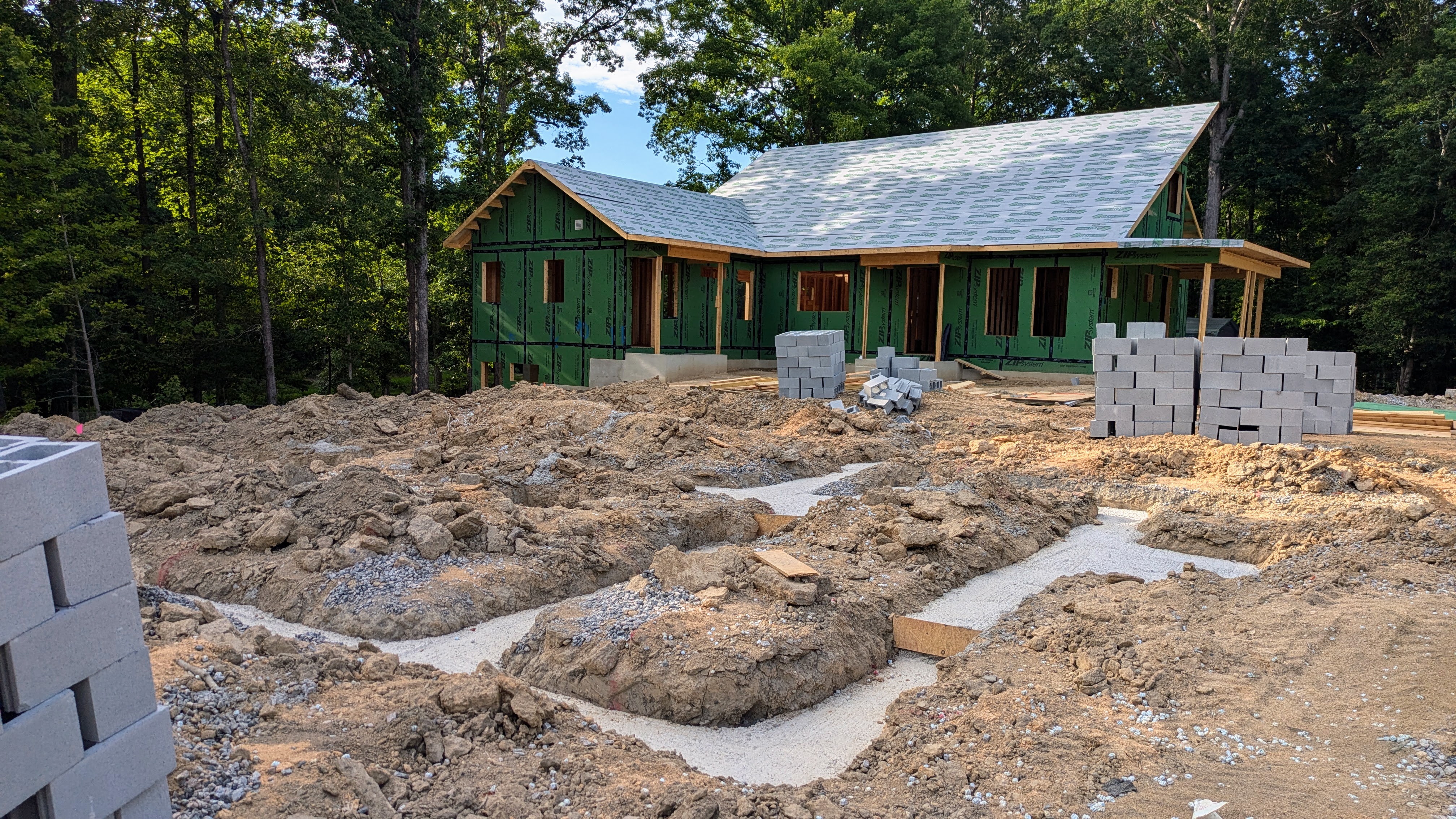 Framed house with Zip system siding and roof sheathing on in the background, concrete foundation and dirt with stacks of blocks for another home foundation in the foreground, surrounded by trees. 