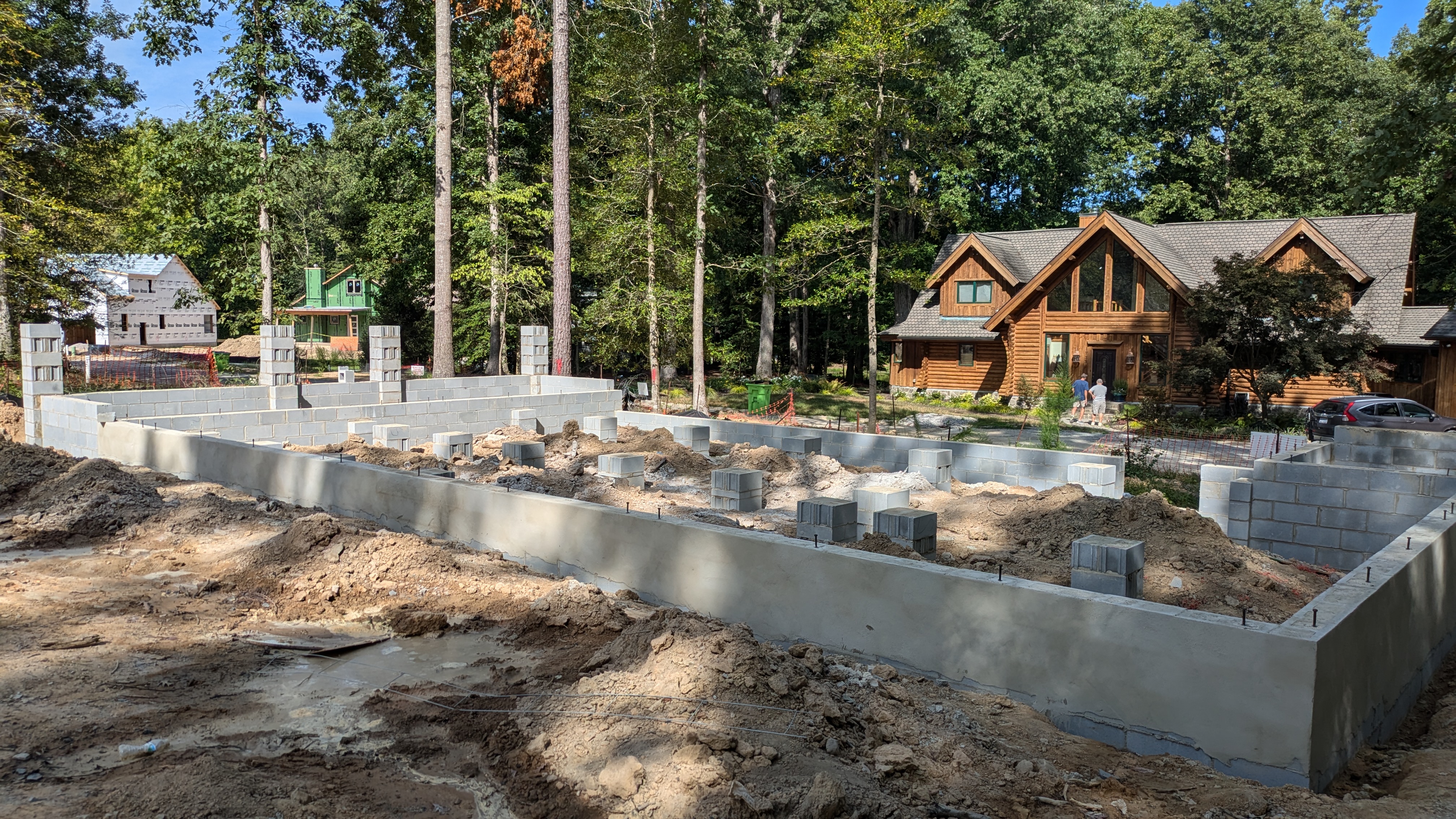 The cement block foundation of a home is shown with pillars in the middle, waiting for the floor joists to be installed. There is a large log cabin in the background.