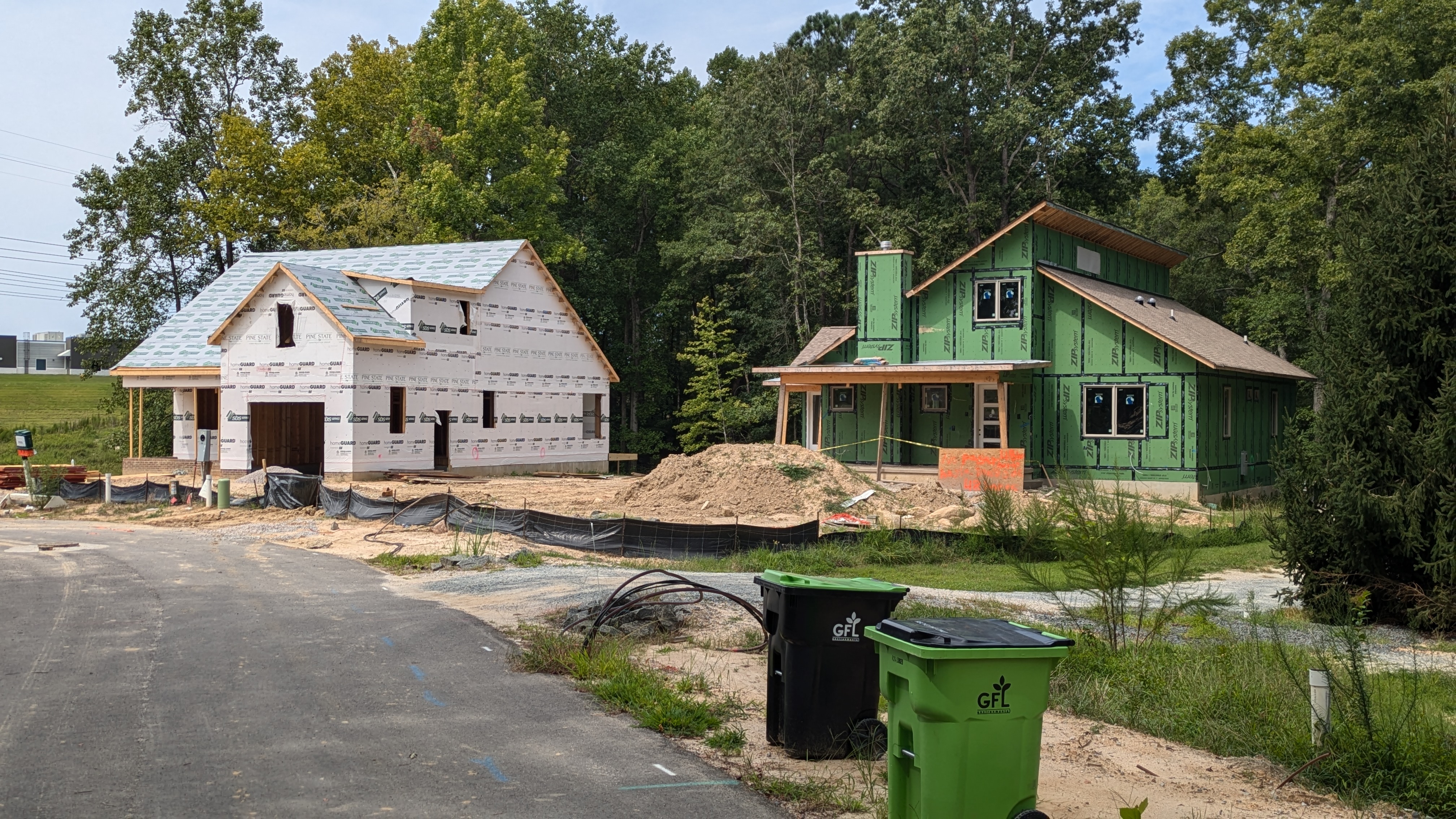 Two houses under construction. One with green sheathing and shingles on the roof and other with white house wrap and roof covering but no shingles. 