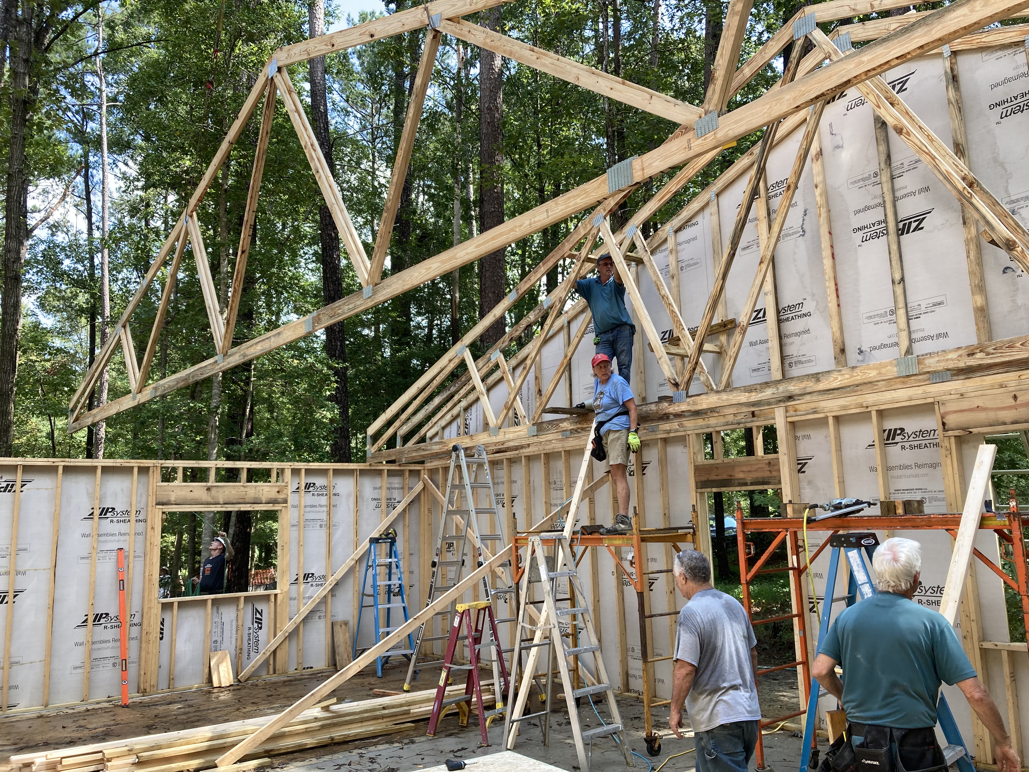 A large roof truss is being lowered onto the framing of a house by a crane with several people guiding it in and getting ready to secure it in place. 