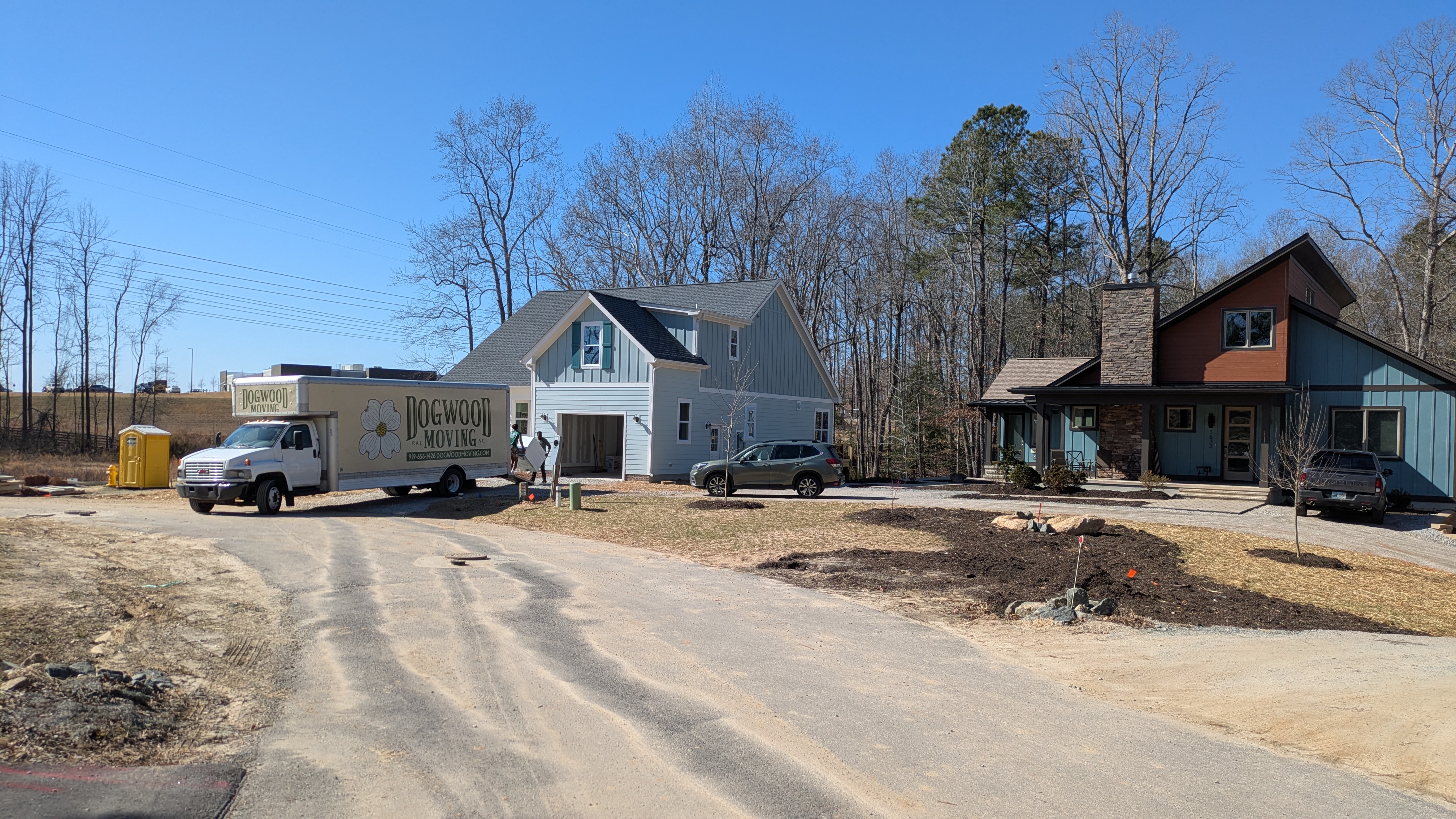 A moving truck back up to a home with movers unloading a dryer. A completed house is to the right with a car in the driveway. 