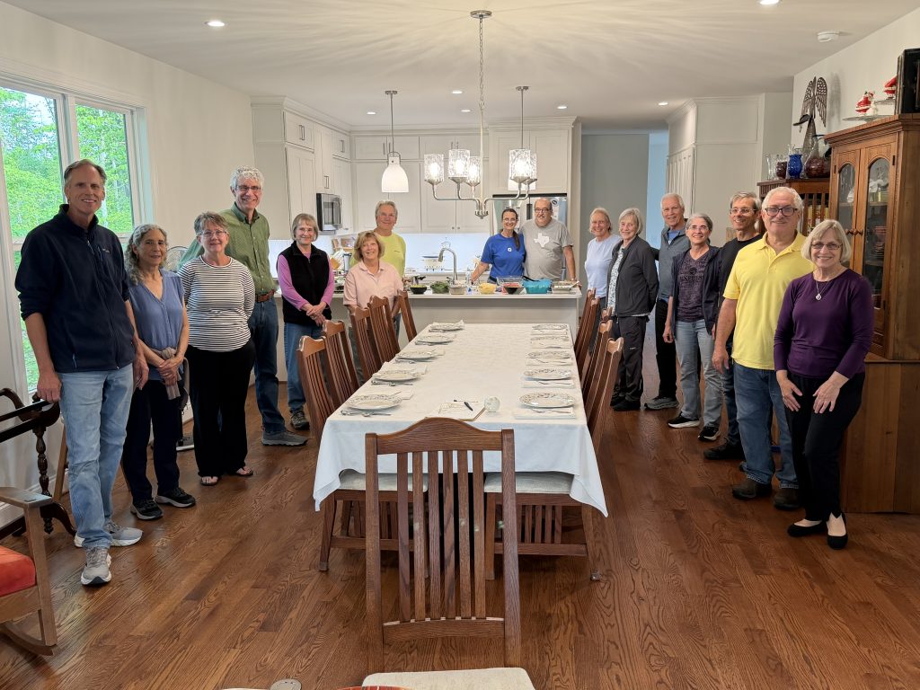 A group of 15 people stand around a large dining room table with a kitchen in the background.